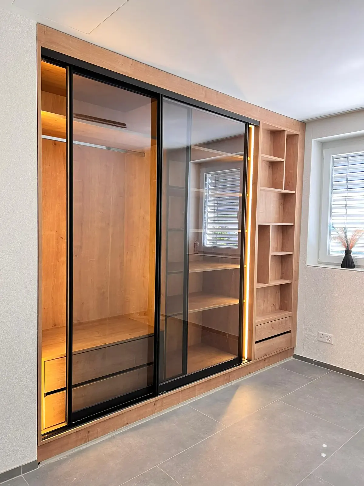Maßgefertigter Glasschrank mit getönten Schiebetüren, schwarzem Rahmen, Holzdekor, integrierter LED-Beleuchtung und offenen Regalfächern.
